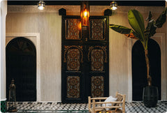 Cream travertine flexible stone veneer applied on Moroccan riad courtyard walls – warm cream surface with soft horizontal grain framing ornate carved black wooden door, lantern pendant lights, banana plant and zellige tile floor