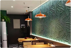 Palm Leaf Relief MCM flexible stone panel as full side wall cladding in a tropical-style restaurant, deep teal-green tone with palm leaf relief pattern dramatically highlighted by horizontal LED strip lighting above, two orange-red vintage enamel dome pendant lights, timber square dining tables, rattan dining chairs and black and white chequerboard floor