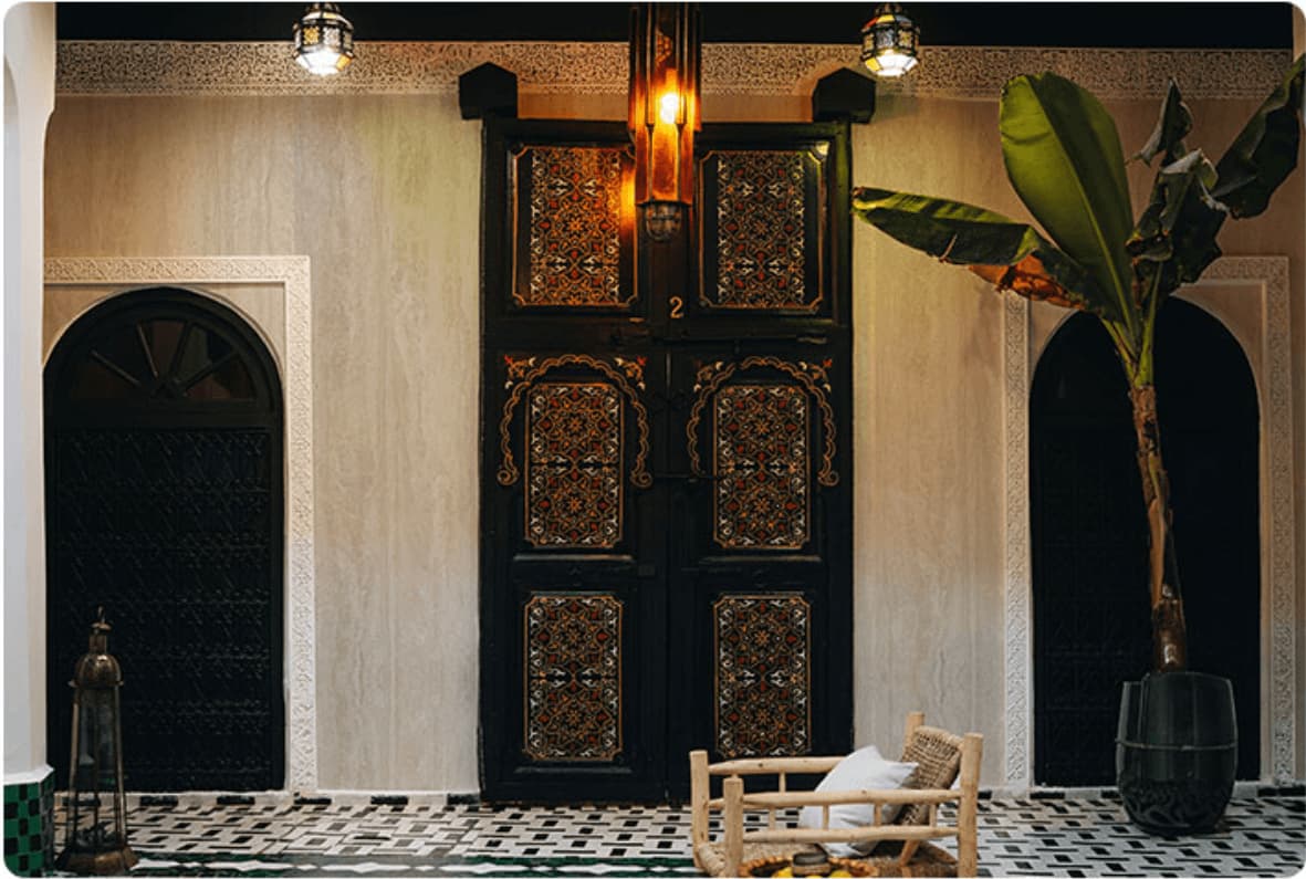 Cream travertine flexible stone veneer applied on Moroccan riad courtyard walls – warm cream surface with soft horizontal grain framing ornate carved black wooden door, lantern pendant lights, banana plant and zellige tile floor