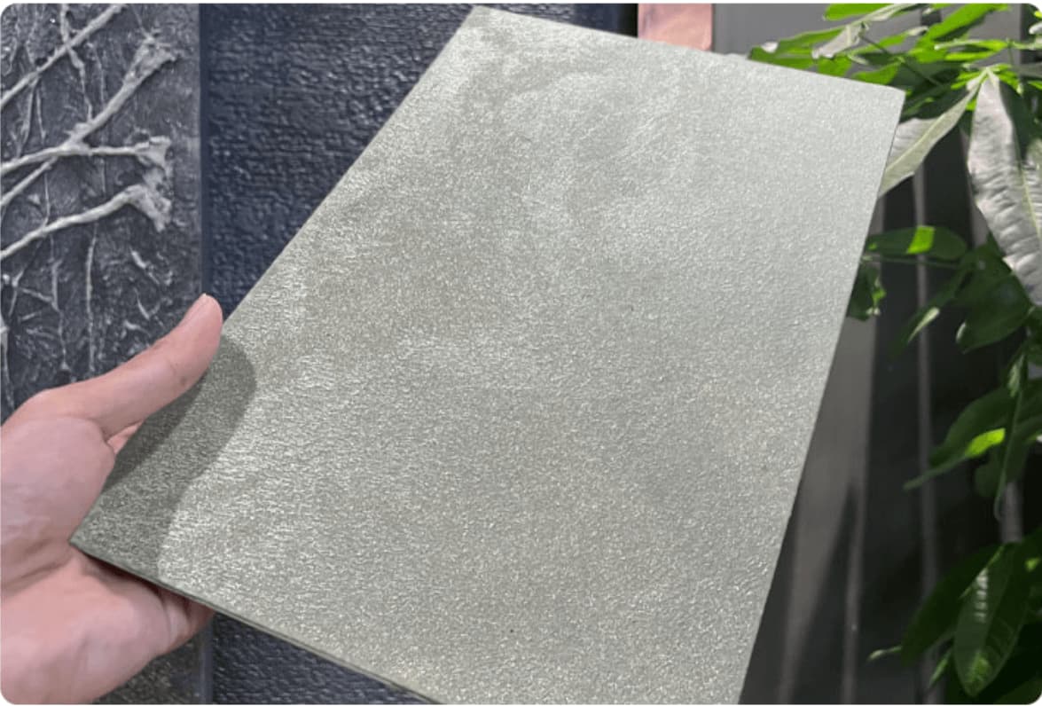 Satin Platinum metallic flexible wall panel – hand-held panel showing cool silver hammered foil surface with fine iridescent texture, outdoor showroom with dark textured stone wall and tropical foliage in background