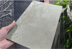 Satin Platinum metallic flexible wall panel – hand-held panel showing cool silver hammered foil surface with fine iridescent texture, outdoor showroom with dark textured stone wall and tropical foliage in background